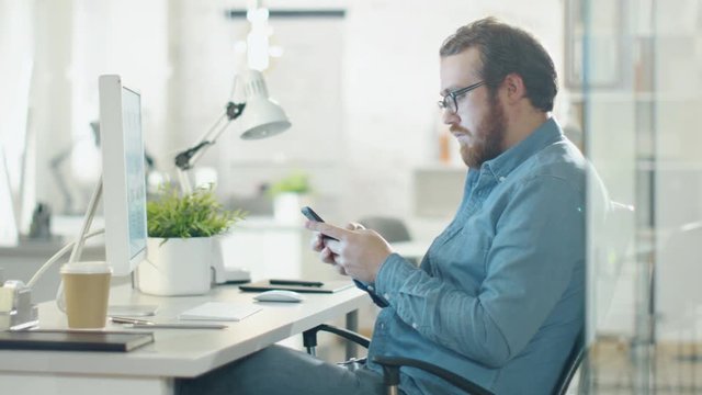 Creative Man Uses Smartphone while Sitting at His Workplace. Office is Light and Modern with Green Plant on the Table. Shot on RED Cinema Camera in 4K (UHD).