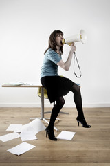 Full length of businesswoman screaming into megaphone while sitting on desk in office
