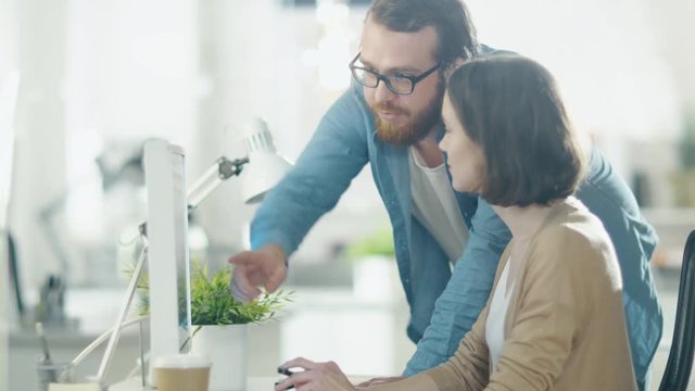 Young Man and Beautiful Woman Discuss Working Issues at Workplace. She Sits at the Table. He Stands Beside Her. They Point at Something on the Screen. Shot on RED Cinema Camera in 4K (UHD).