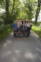 Full length portrait of couple with three kids sitting on back of trailer on country lane
