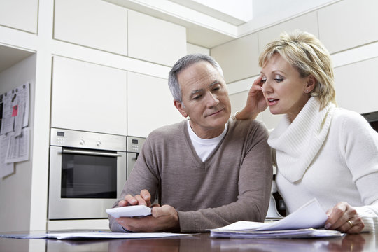 Middle Aged Couple Making Calculations Using Calculator At Kitchen Table