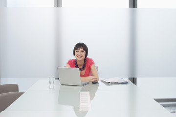 Smiling thoughtful young woman sitting with laptop in modern cubicle at office