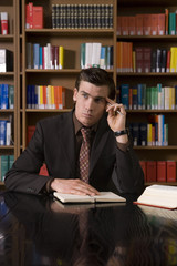 Thoughtful young man in formals with book and pen at desk in library