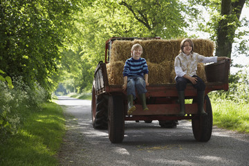 Full length portrait of two young boys sitting on back of trailer on country lane