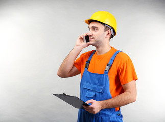 Handsome warehouse worker talking by mobile phone while holding clipboard, on light background