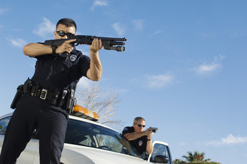 Low angle view of two police officers aiming shotguns by patrol car
