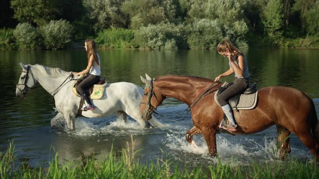 CLOSE UP: Two young girls riding horses along grassy river bank