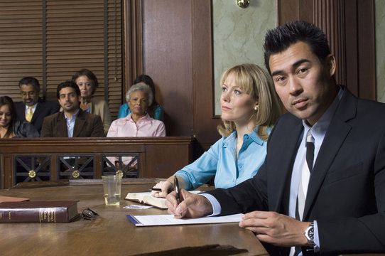 Portrait Of A Male Defense Lawyer Sitting With Client In Courtroom