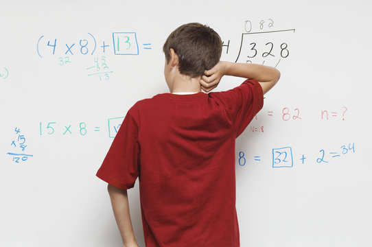 Rear view of a teenage boy scratching head against white board in classroom