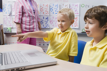 Schoolboy pointing at laptop while sitting with classmate in classroom