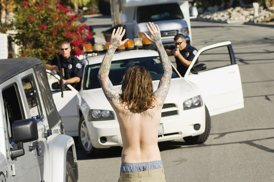 Rear view of a shirtless young man surrendering with two police officers aiming guns
