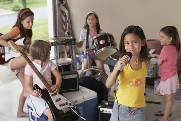 Portrait of cute young girl singing into microphone with friends playing musical instrument in...