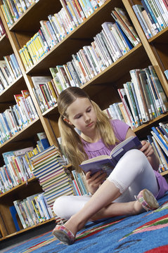 Teenage girl reading book while sitting on floor in library