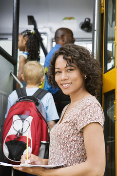 Portrait of beautiful female teacher loading elementary students on school bus