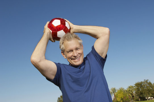 Portrait Of A Happy Senior Man Throwing Soccer Ball Against Blue Sky