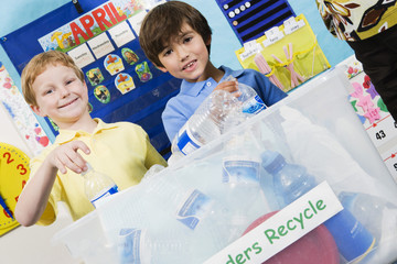 Two elementary students throwing plastic bottles in recycling container at classroom
