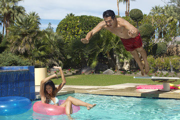 Happy young man jumping into swimming pool over woman on inflatable raft