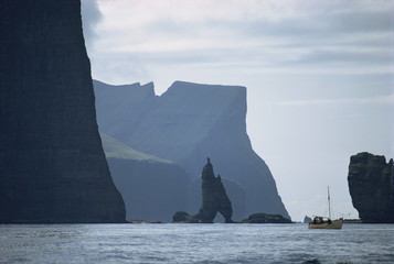 Small fishing boat before a towering cliff coastline, Faroe Islands, Denmark, Atlantic