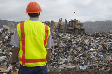 Rear view of a worker watching digger move waste at landfill site