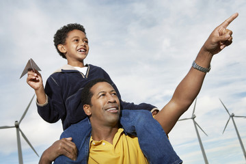 Little boy with paper plane sitting on father's shoulders at wind farm
