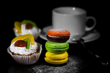 Brightly lit foreground with cakes and vivid colored macaroons in stack gradually moving to dark background with wihte cup for coffee and spoon on saucer/shallow depth of field
