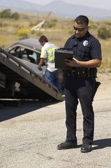 Police officer writing notes with tow truck driver lifting crashed car in background