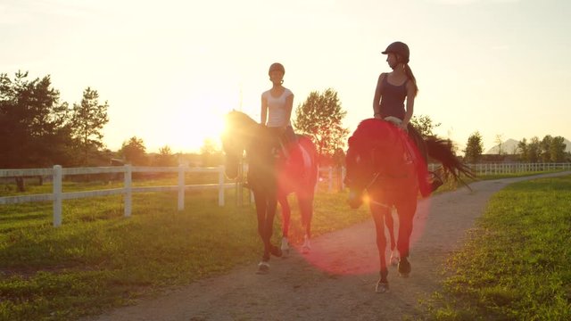 SLOW MOTION: Two smiling friends horseback riding brown horses at golden sunset