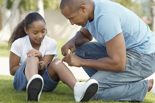 African American father sticking adhesive plaster to daughter's knee at park