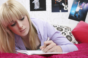 Beautiful young female student writing in book on bed