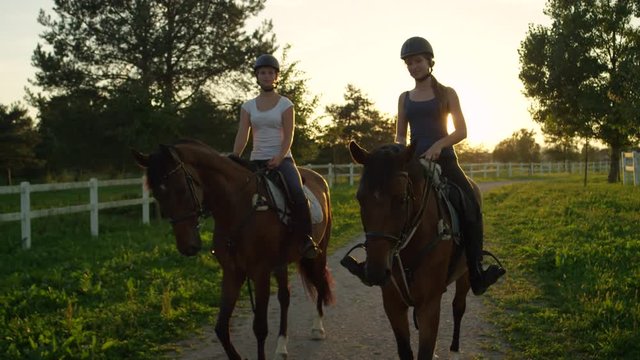 SLOW MOTION: Two happy friends horseback riding brown horses at golden sunset