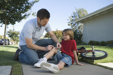 Full length of a father putting plaster on son's knee by bicycle on pathway