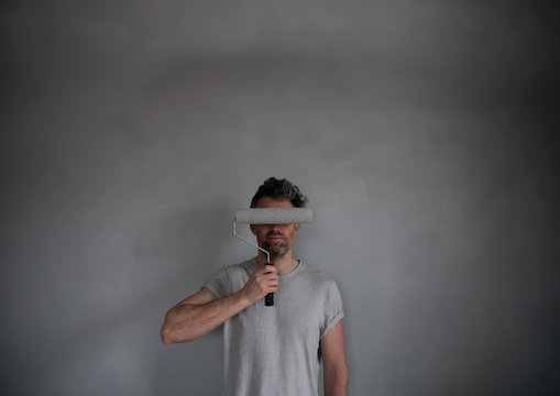 Man covering his eyes with paint roller while standing against gray wall