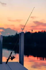 Papier Peint photo Pêcher Une canne à pêche avec des appâts pour grenouilles repose sur un quai au coucher du soleil sur le lac.