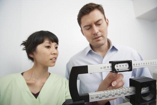 An Asian Female Nurse Adjusting Weighing Scale For Man At Clinic