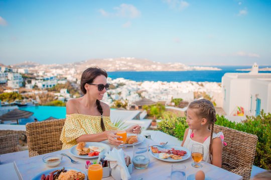 Family Having Breakfast At Outdoor Cafe With Amazing View On Mykonos Town. Adorable Girl And Mom Drinking Fresh Juice And Eating Croissant On Luxury Hotel Terrace