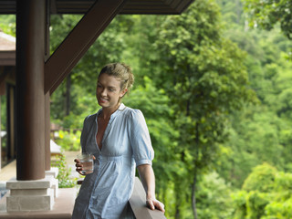 Fototapeta premium Smiling young woman with a glass of water in veranda against blurred trees