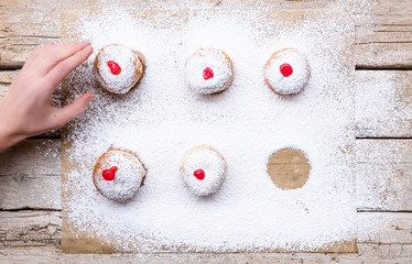 Fresh donuts (sufgania) with red jelly jam on a sugar powder and wooden background. Hand of woman taking donut from bakery paper. Hanukkah holiday celebration and traditional jewish sweet