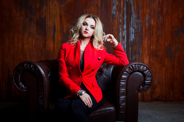 Young and attractive blond woman in red jacket sits in leather armchair, background grunge rusty wall