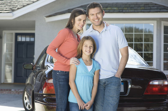 Portrait Of A Happy Couple With Daughter Standing Against Car And House