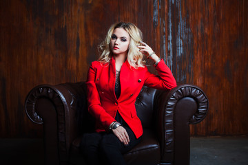 Young and attractive blond woman in red jacket sits in leather armchair, background grunge rusty wall