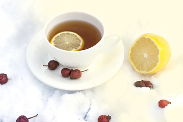 white mug of tea on a saucer with a lemon and wild rose on snow