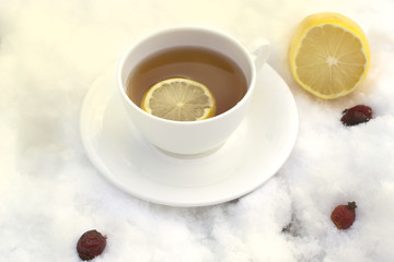 white mug of tea on a saucer with a lemon and wild rose on snow
