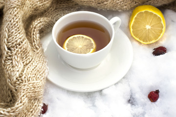 white mug of tea wrapped in a knitted scarf on a saucer, lemon a