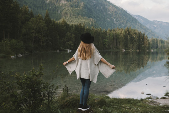Blonde young adult woman wearing a poncho and a hat enjoying a morning in the forest at a mountain lake