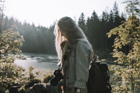 Portrait of a Blonde young adult woman photographer wearing a jeans jacket and a backpack standing in a forest in front of a lake at sunrise
