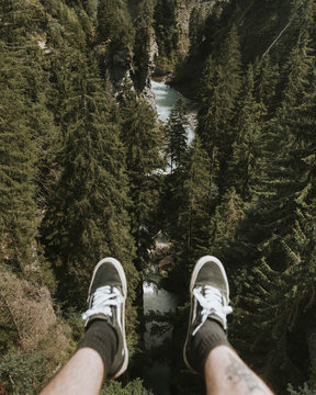 Legs of Adult male with a tattoo wearing black shoes sitting and overlooking a river and pine trees at bright daylight