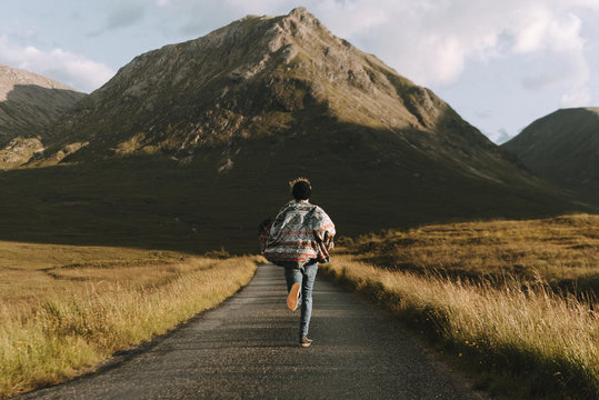 Adventurous Young adult male wearing jeans and a colorful flanel shirt running down a road towards a mountain at sunset