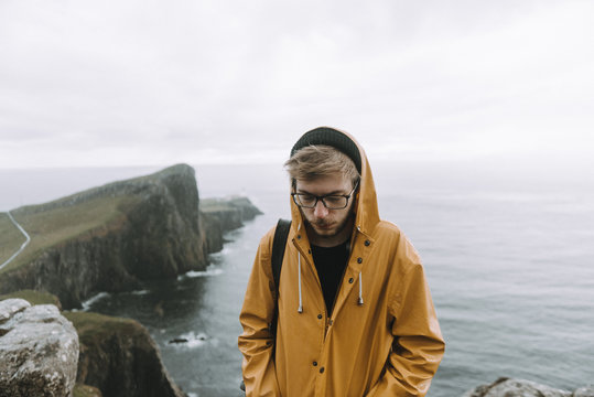 Portrait of a young male wearing glasses and a yellow raincoat standing in the rain with the coastline cliffs and the ocean in the background