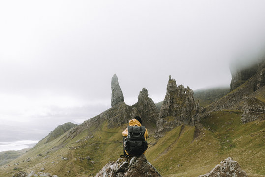 Young adult man wearing a yellow coat and black backpack sitting in front of the old man of storr volcanic stone formation panorama with a dramatic and hazy sky