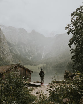 Young male standing by a foggy moody lake near a boat house and forest with mountains in the background